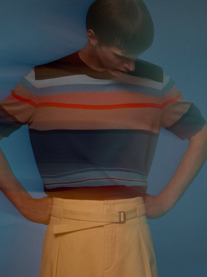 Model Kris Wardak posing with hands on hips wearing a multi-striped knit shirt and cream belted trousers against a blue backdrop.