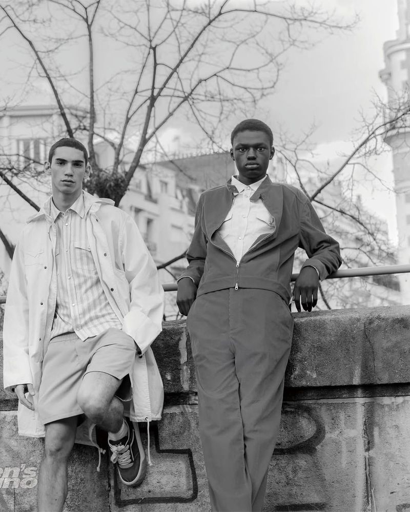 Two male models posing outdoors in tailored designer menswear against a concrete ledge and bare trees in black and white.