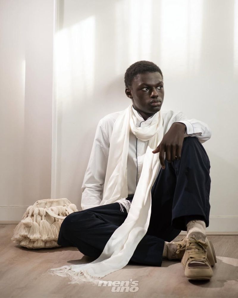 A seated Black male model wearing a white collared shirt, long draped white scarf, and dark navy trousers on a wooden floor.