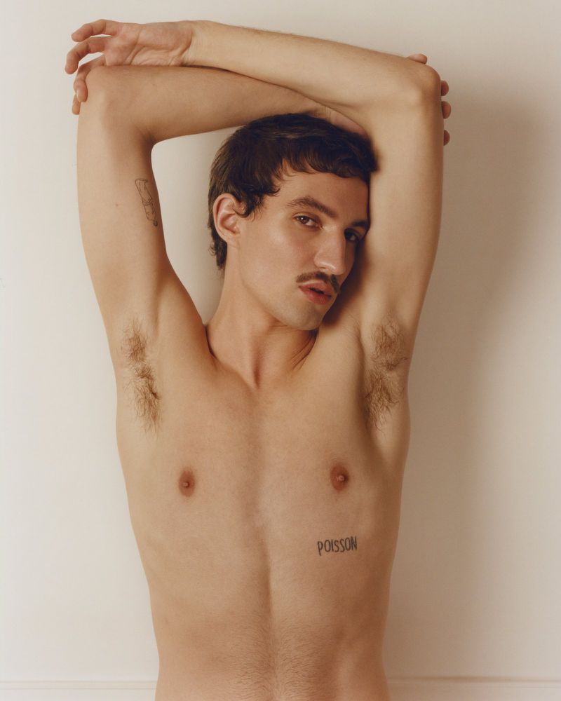 Shirtless young man with a mustache and tattoos posing with arms raised behind his head against a plain off-white wall.