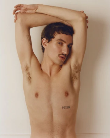 Shirtless young man with a mustache and tattoos posing with arms raised behind his head against a plain off-white wall.