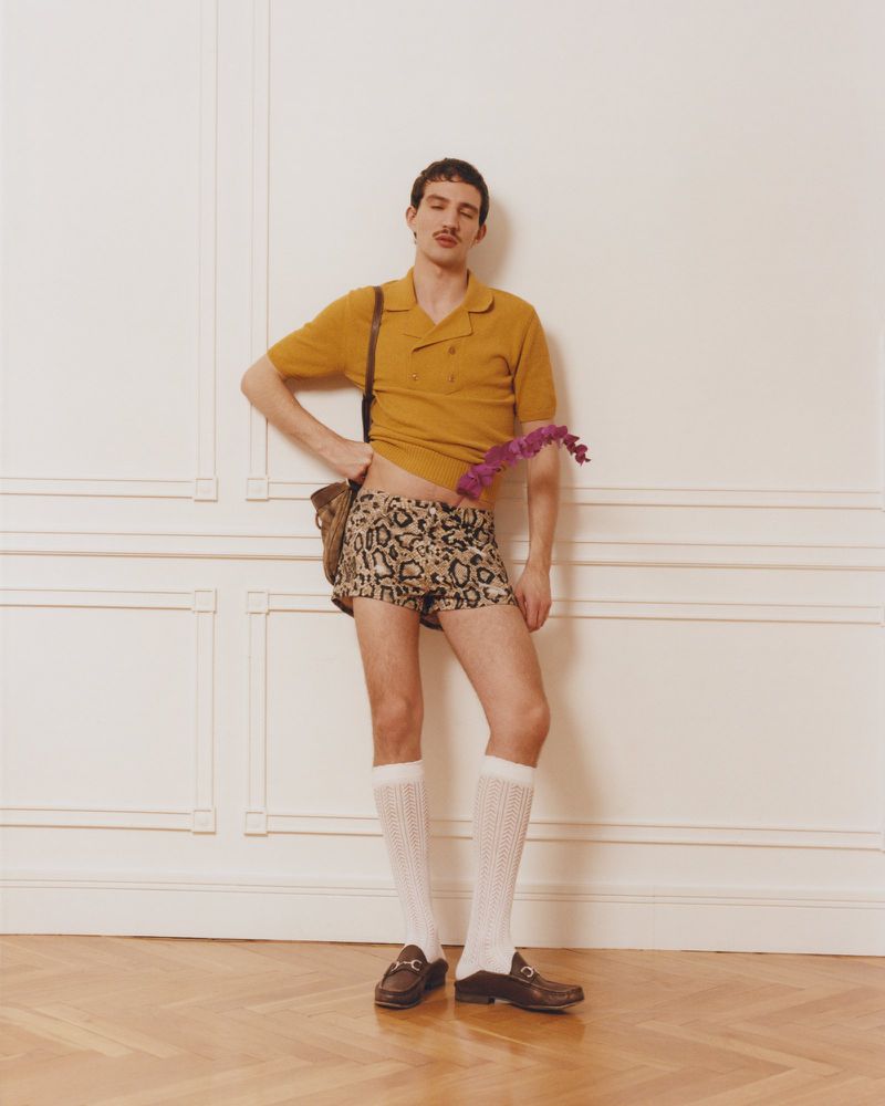 Man in a mustard knit polo and leopard print shorts posing in white knee-high socks and loafers against a white paneled wall.