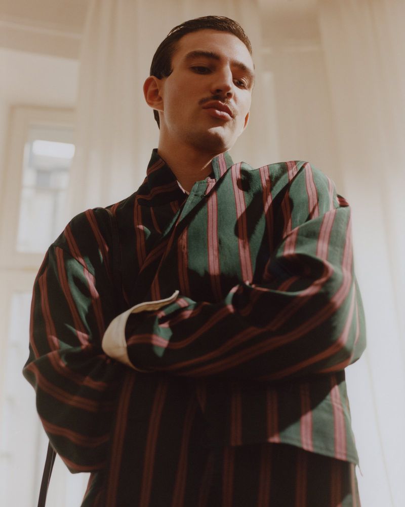 Low-angle portrait of a young man with a mustache wearing a green and burgundy vertical striped jacket with arms crossed.