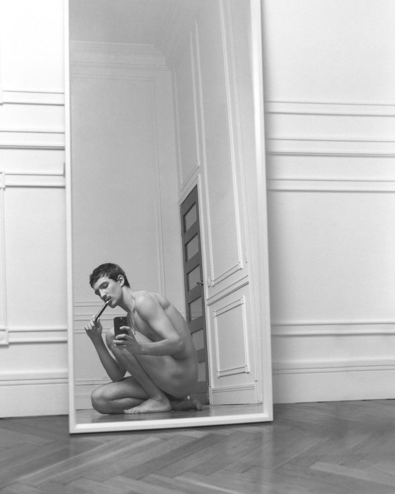 Black and white reflection of a young man crouching on the floor while brushing his teeth and taking a photo in a full-length mirror.