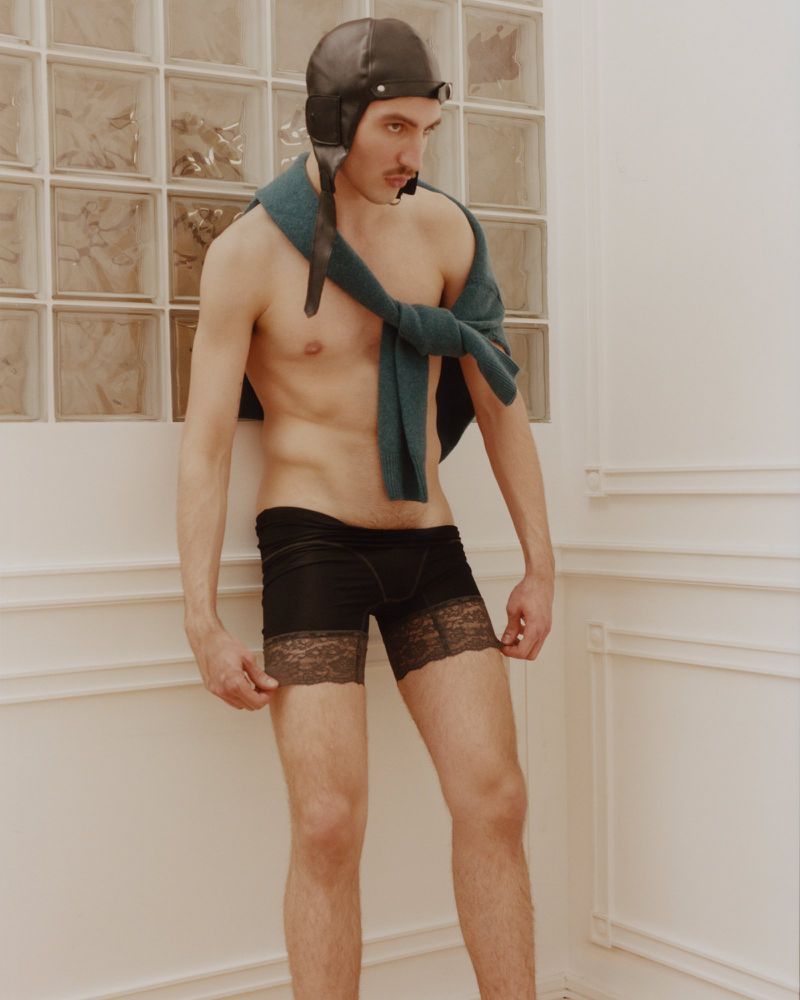 Shirtless man wearing black lace-trimmed boxer shorts, a draped teal sweater, and a leather aviator cap against an interior wall.