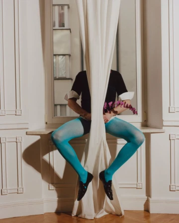 Person seated on a windowsill behind a white curtain wearing bright blue tights, black shoes, and holding purple flowers.