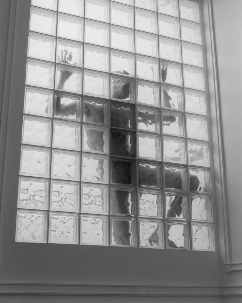 Black and white photograph of a man's silhouette visible behind a tall grid of textured glass blocks with hands pressed on the surface.