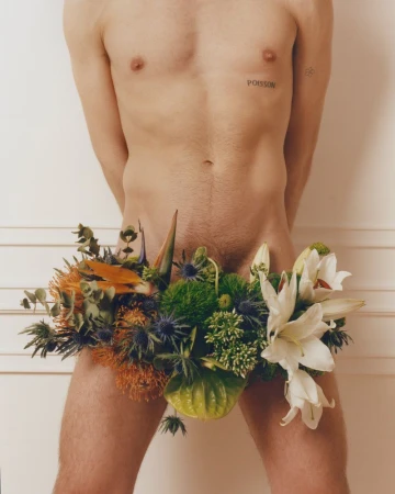 Close-up of a shirtless man's torso with a large arrangement of flowers and greenery arranged over his pelvic area against a light wall.