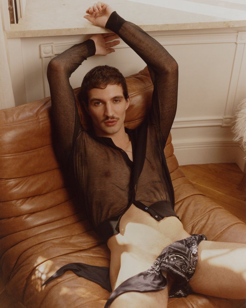 A man reclining on a ribbed brown leather lounge chair wearing a sheer black shirt and a patterned bandana tied at the waist.