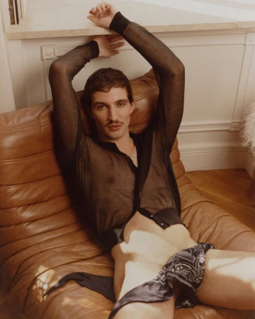 A man reclining on a ribbed brown leather lounge chair wearing a sheer black shirt and a patterned bandana tied at the waist.