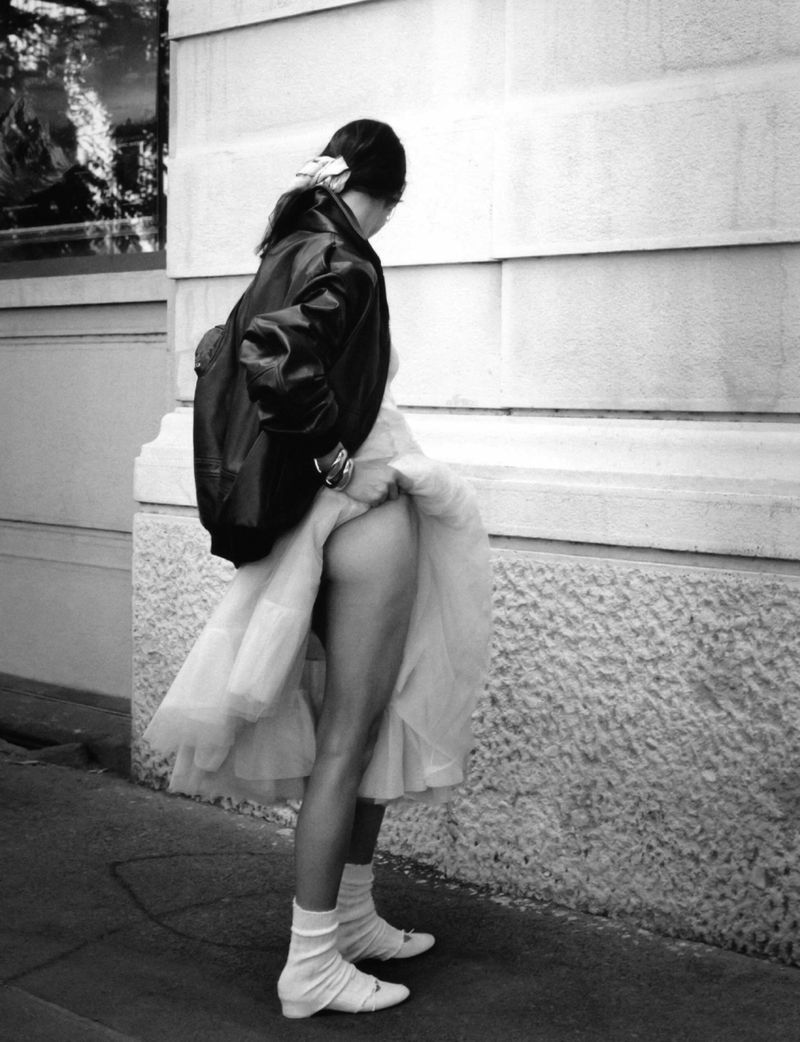 Black and white photo of Vivienne Rohner lifting a white tulle skirt, wearing a black leather jacket on a sidewalk.