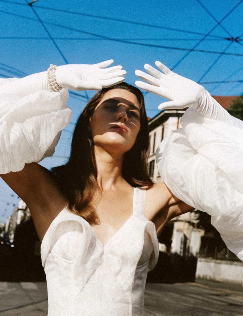 Model Vivienne Rohner in a white corset dress with voluminous sleeves and white gloves raising her hands under a blue sky.