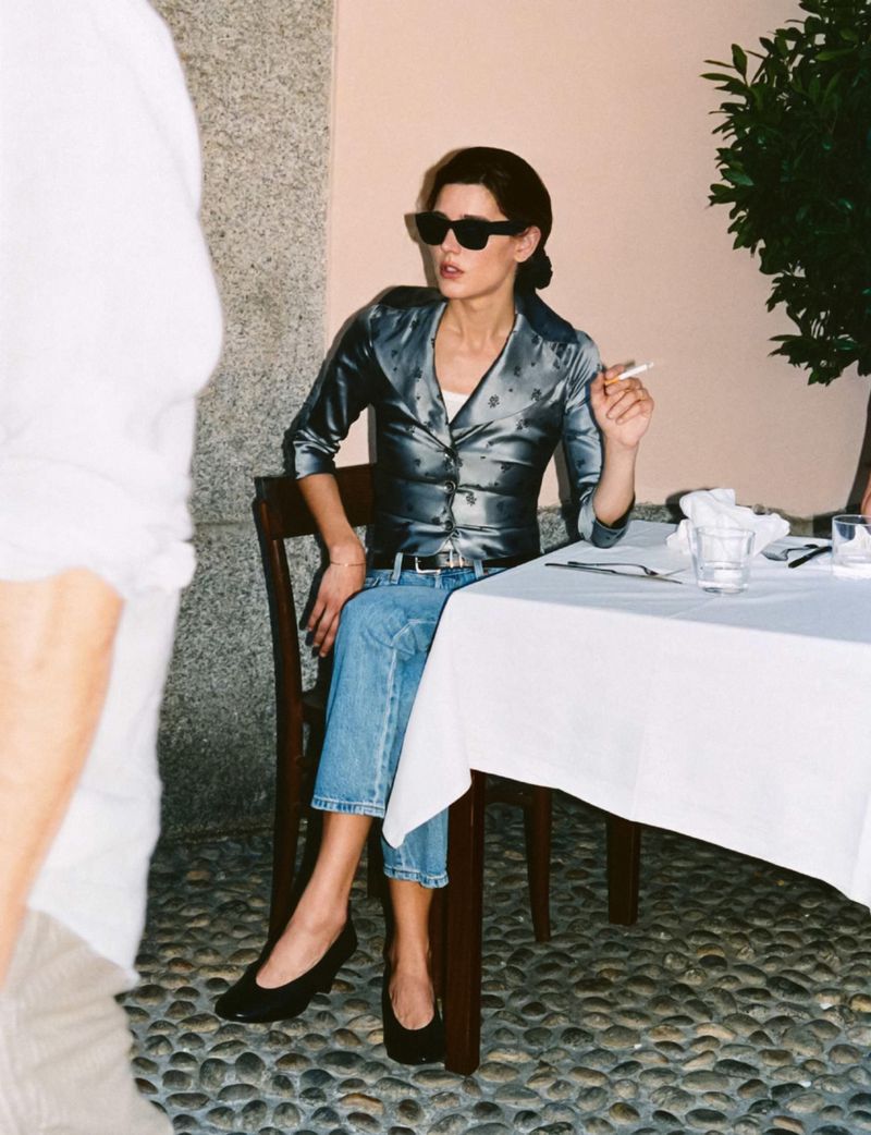 Model Vivienne Rohner seated at an outdoor restaurant table wearing sunglasses, a metallic shirt, and blue jeans holding a cigarette.
