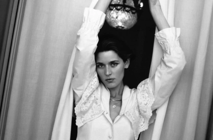 Black and white portrait of model Vivienne Rohner with arms raised between hanging curtains wearing a white lace-trimmed blouse.