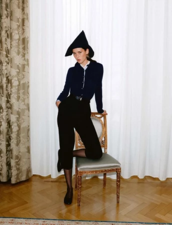 Model Vivienne Rohner in a pointed black hat and dark navy cardigan posing with one knee on an antique chair against white curtains.