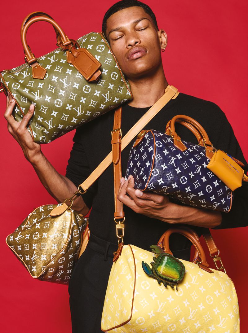 Fashion shot of a black male model with various Louis Vuitton monogram bags in a studio setting against a red backdrop.