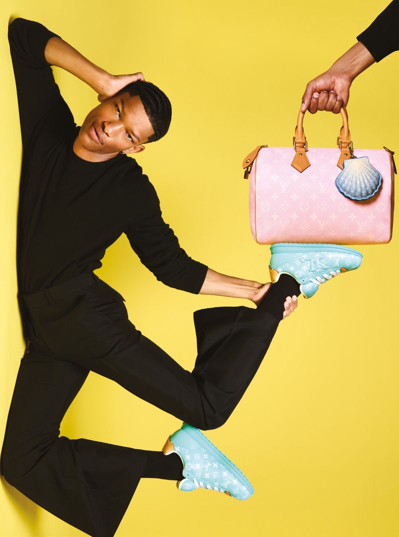 Person in black outfit balancing on shoes with a Louis Vuitton bag, against a yellow backdrop in a studio setting.