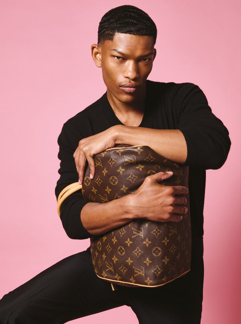 Elegant adult male poses in black with Louis Vuitton bag against pink backdrop, showcasing fashion and luxury in a portrait style.