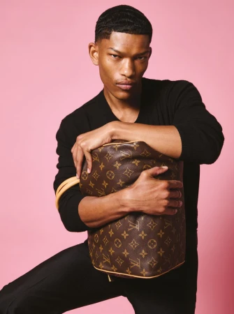 Elegant adult male poses in black with Louis Vuitton bag against pink backdrop, showcasing fashion and luxury in a portrait style.