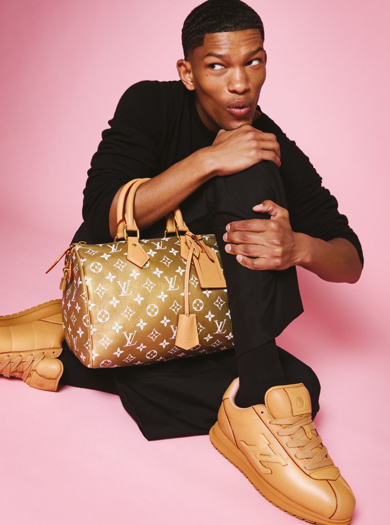 Stylish African-American man poses with a Louis Vuitton handbag and tan sneakers against a pink backdrop, showcasing a modern fashion look.