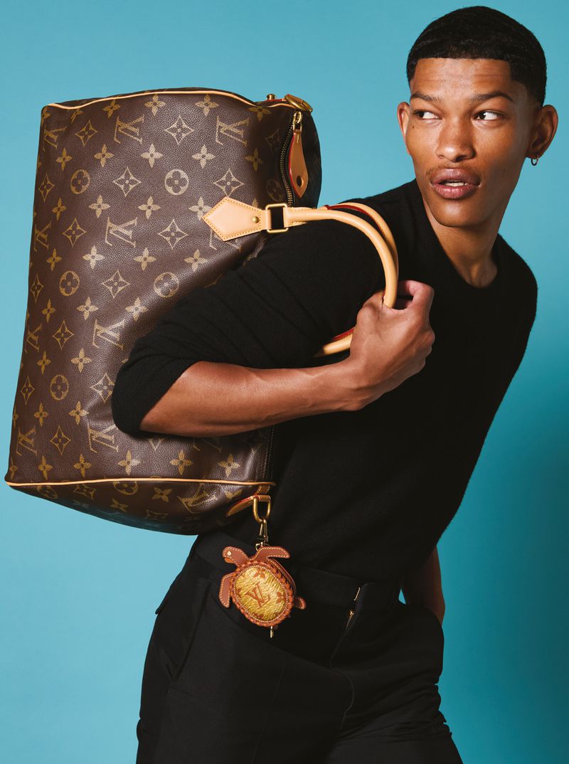 Young adult carries a Louis Vuitton duffel bag. He wears a black shirt and trousers, posing against a blue backdrop in a studio.