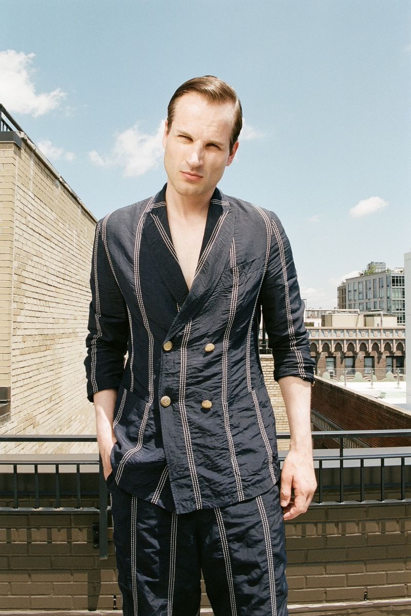 David Koch wearing a navy striped double-breasted suit without a shirt on an urban rooftop under direct sunlight.