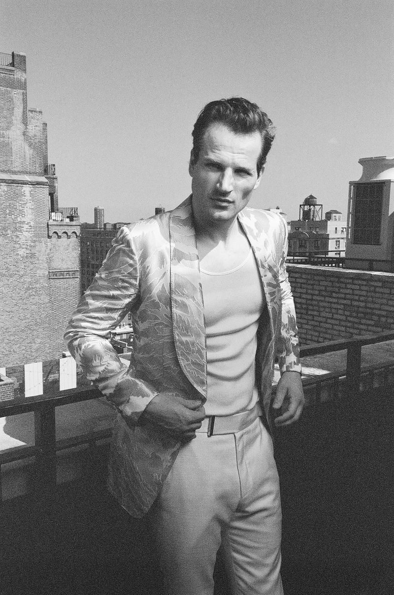 Model David Koch poses on a city rooftop wearing a patterned shawl-lapel blazer and light trousers in a black and white fashion editorial.