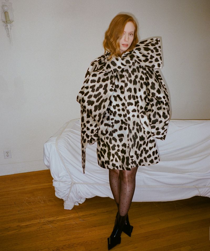 Hannah Einbinder posing in a voluminous leopard-print cape dress with black tights and boots in front of a draped sofa.