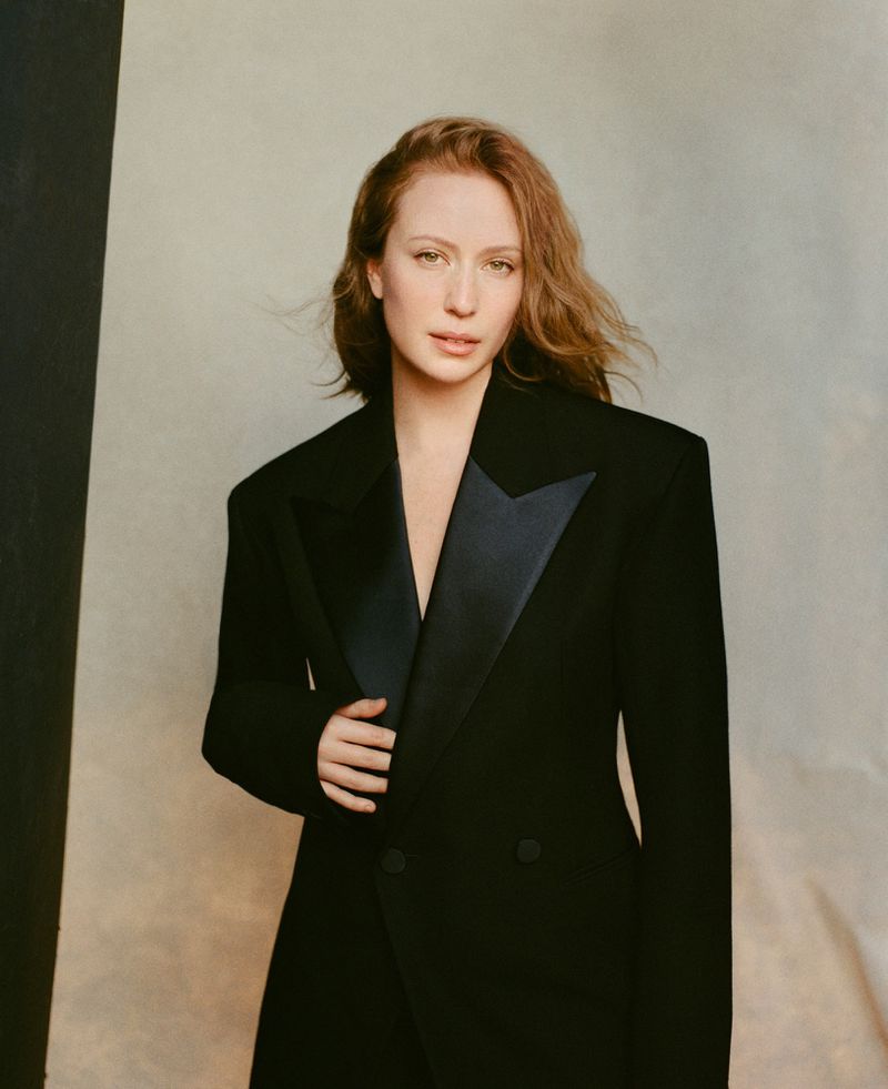Hannah Einbinder posing in a tailored black tuxedo blazer with satin lapels against a neutral studio backdrop.