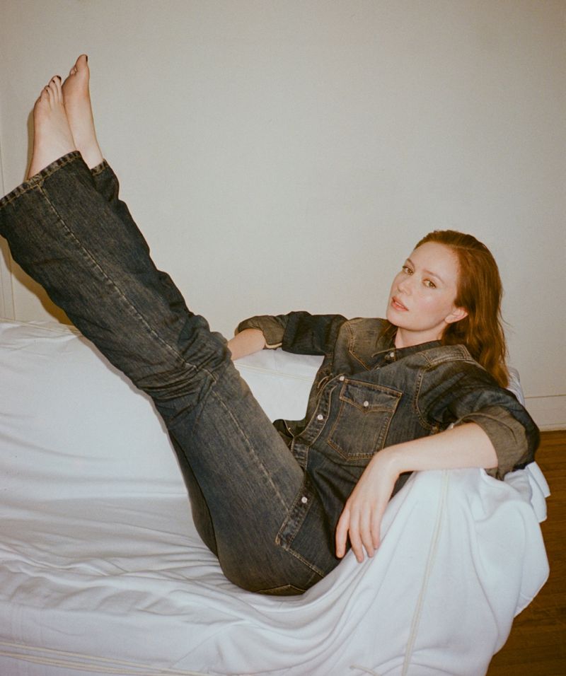 Hannah Einbinder reclining on a white-draped couch with bare feet raised, wearing a dark wash denim shirt and jeans.