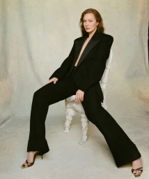Hannah Einbinder seated on a wrapped white chair wearing an open black tuxedo suit with matching trousers and strappy heels.