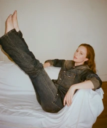 Hannah Einbinder reclining on a white-draped couch with bare feet raised, wearing a dark wash denim shirt and jeans.