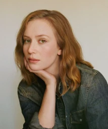 Hannah Einbinder resting her chin in her hand while wearing a dark denim button-up shirt against a pale studio background.