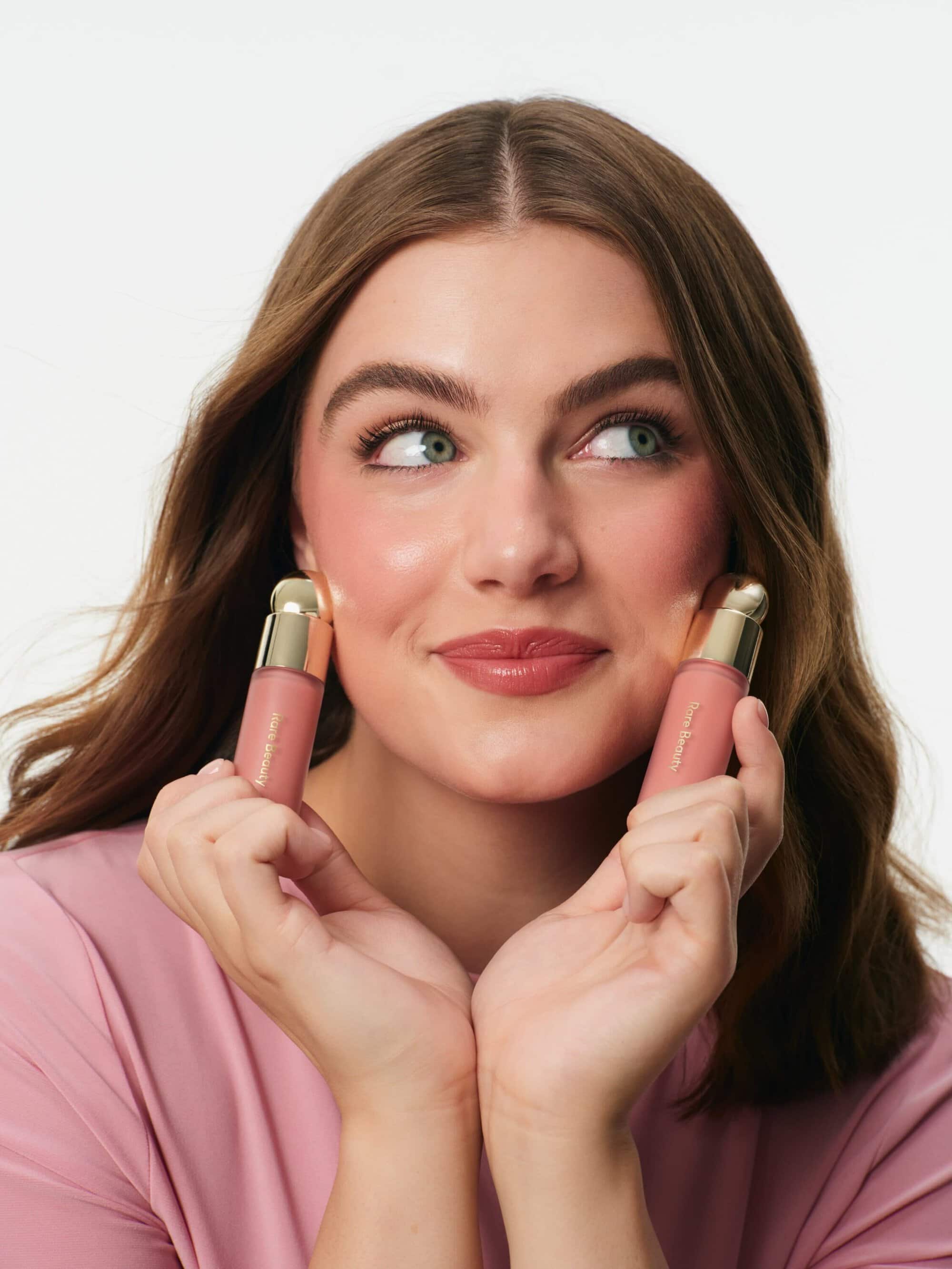 Model Ella Bright smiling and holding two pink liquid blush bottles to her face against a bright studio background.