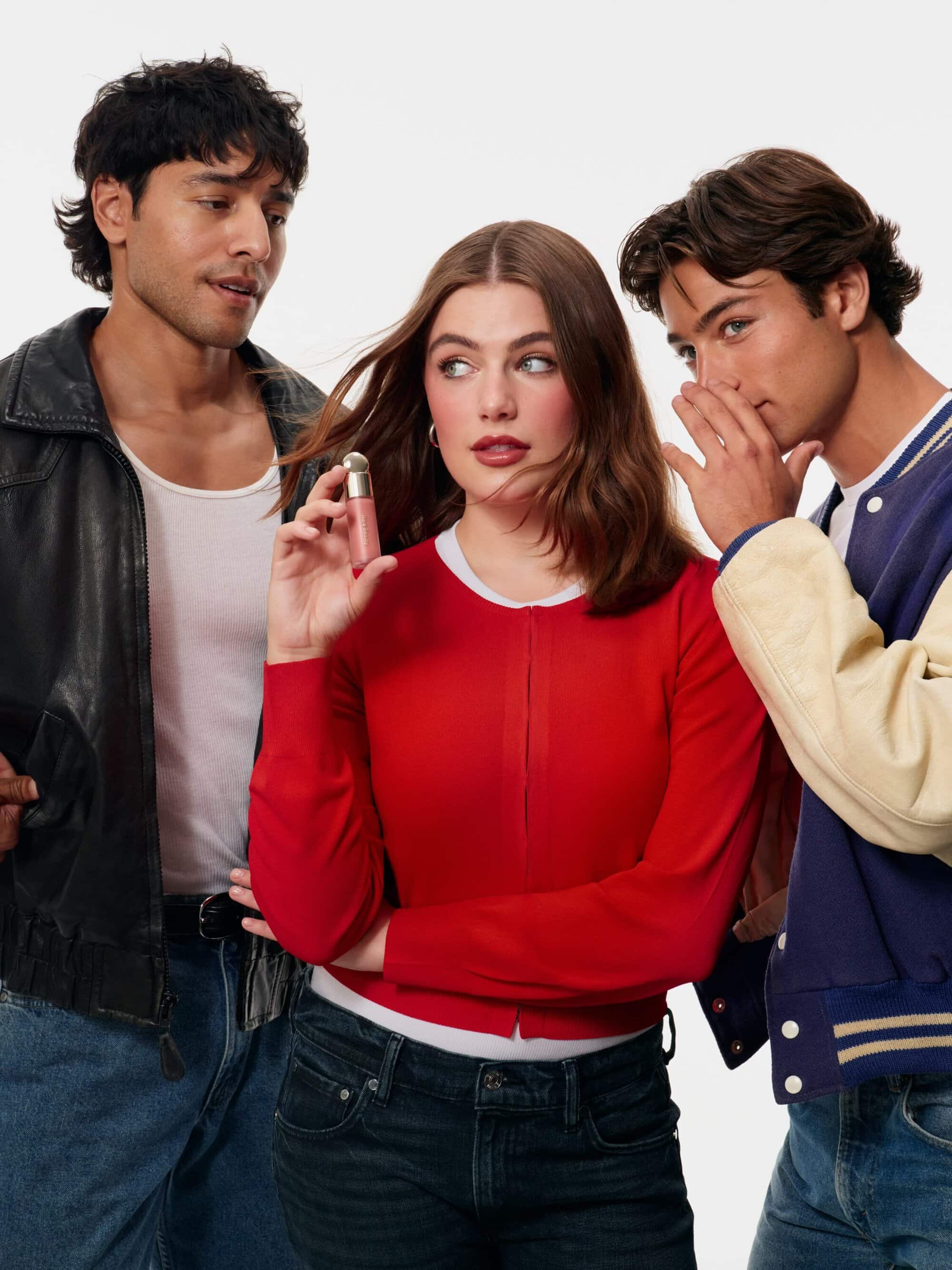 Model Ella Bright in a red cardigan holding Rare Beauty liquid blush flanked by two men against a white seamless background.