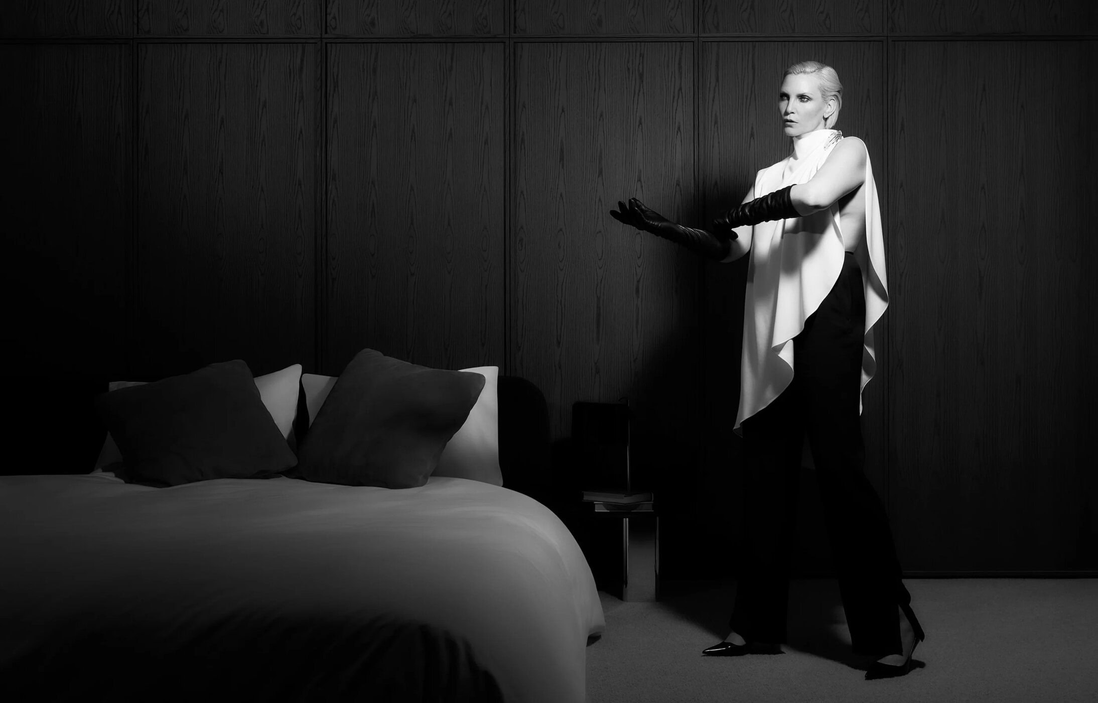 Nadja Auermann standing in a draped white top and long black gloves beside a bed in a dark wood-paneled room.