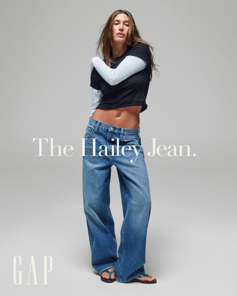 Hailey Bieber wearing baggy blue denim jeans and a cropped layered t-shirt against a light grey studio background.