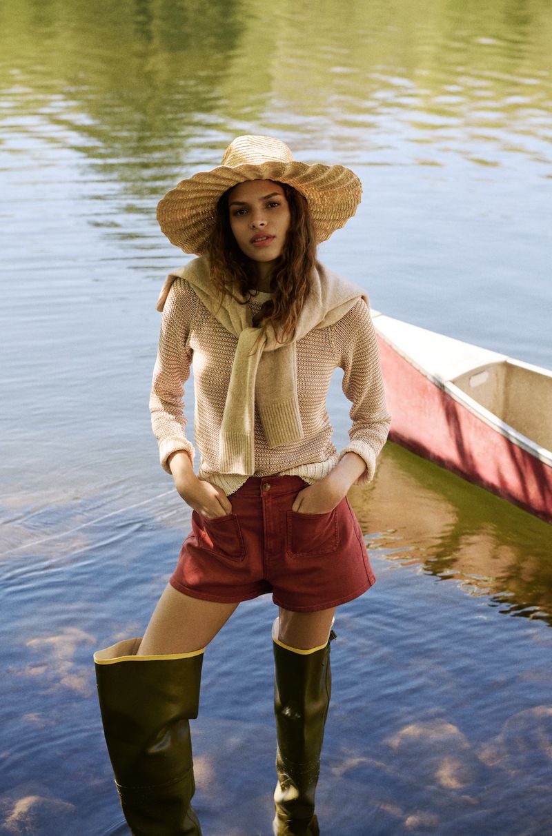 Model in a wide-brim straw hat, beige knit sweater, terracotta shorts, and olive waders standing in lake water by a canoe.