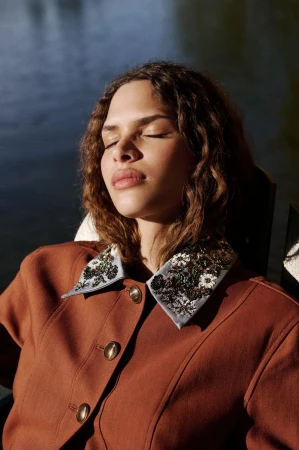 Model Angel Galego with eyes closed wearing a rust brown jacket with an embellished floral collar in direct sunlight.