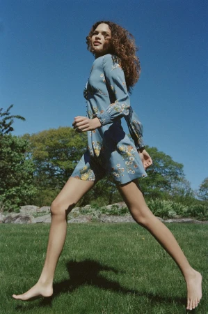 Model with long curly hair wearing a blue floral dress leaping barefoot across a grassy lawn under a clear sky.
