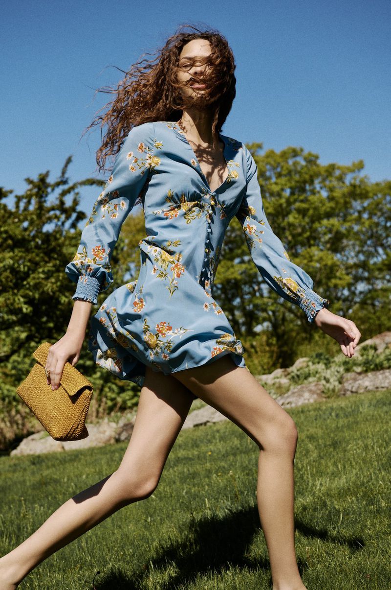 Model running across a green lawn wearing a light blue floral long-sleeve mini dress and carrying a yellow woven clutch.