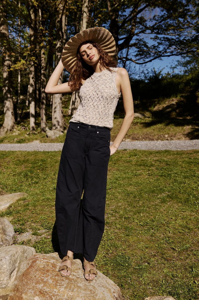 Model wearing a wide-brim straw hat, textured sleeveless knit top, and wide-leg dark trousers standing on a rock outdoors.