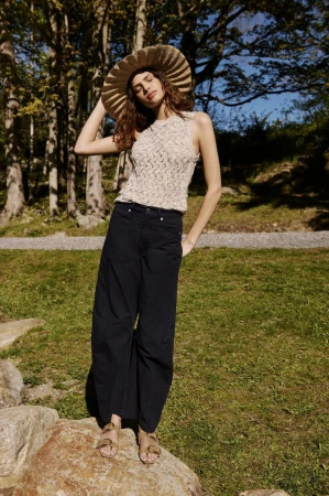 Model wearing a wide-brim straw hat, textured sleeveless knit top, and wide-leg dark trousers standing on a rock outdoors.