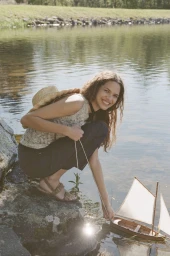 Model crouching on a rocky lake shore in a sleeveless top and dark trousers, guiding a small wooden sailboat in the water.