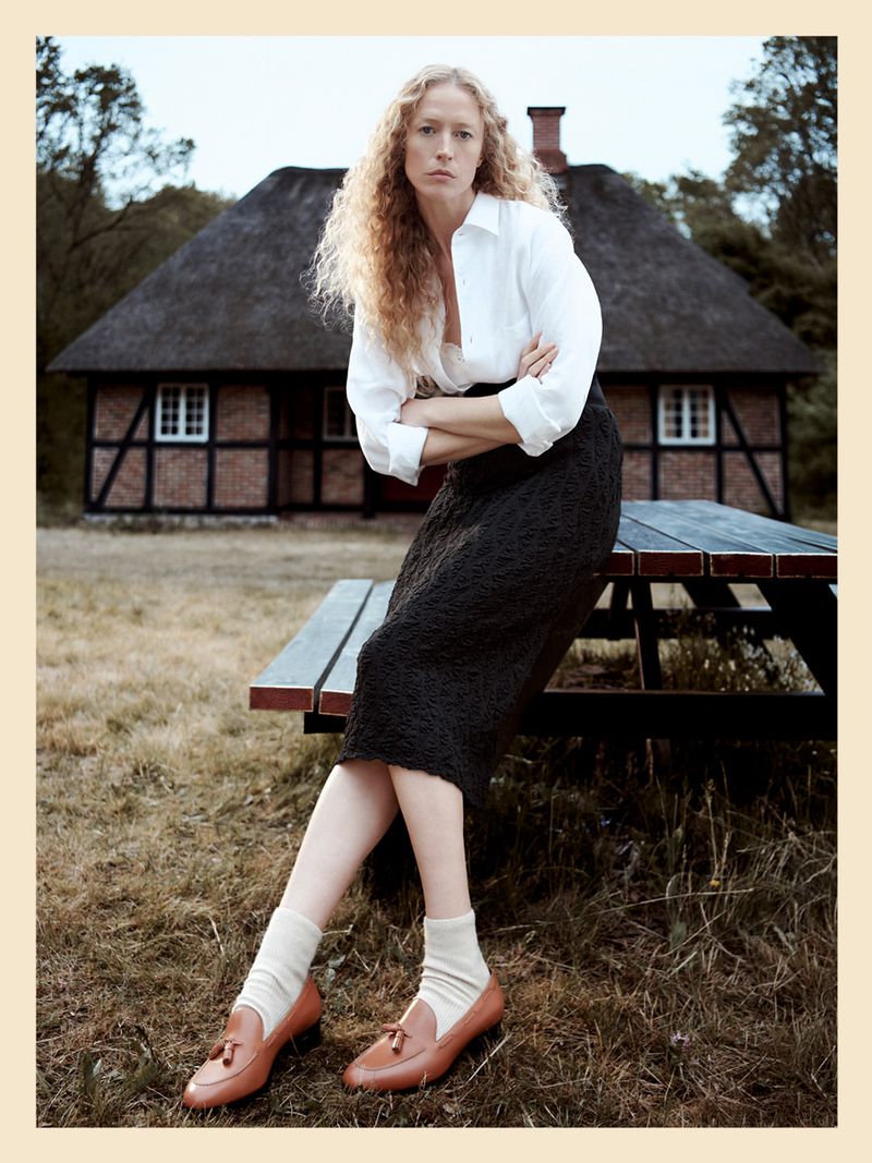Raquel Zimmermann sitting on a wooden picnic table wearing a white shirt, textured midi skirt, and tan loafers in front of a cottage.