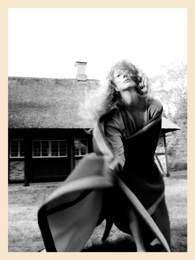 Monochrome portrait of Raquel Zimmermann twirling in a woolen blanket outdoors before a thatched-roof house.