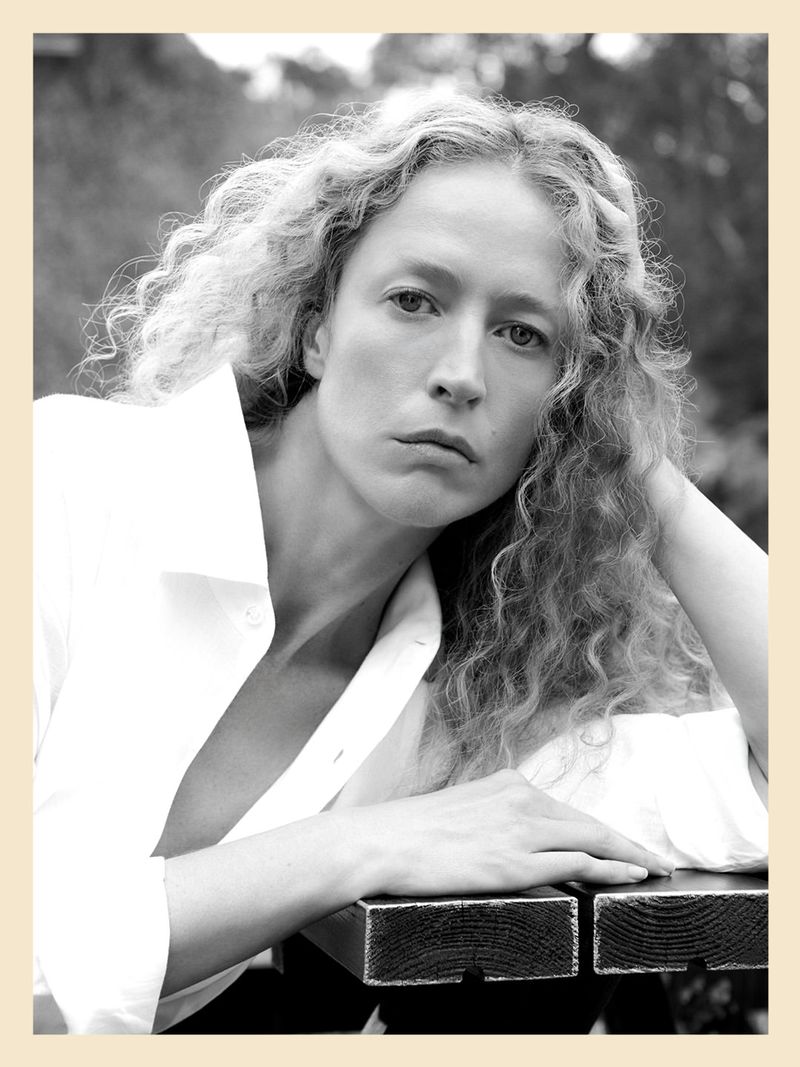 Black and white portrait of Raquel Zimmermann with curly hair resting her hand on her head, wearing an unbuttoned white shirt.