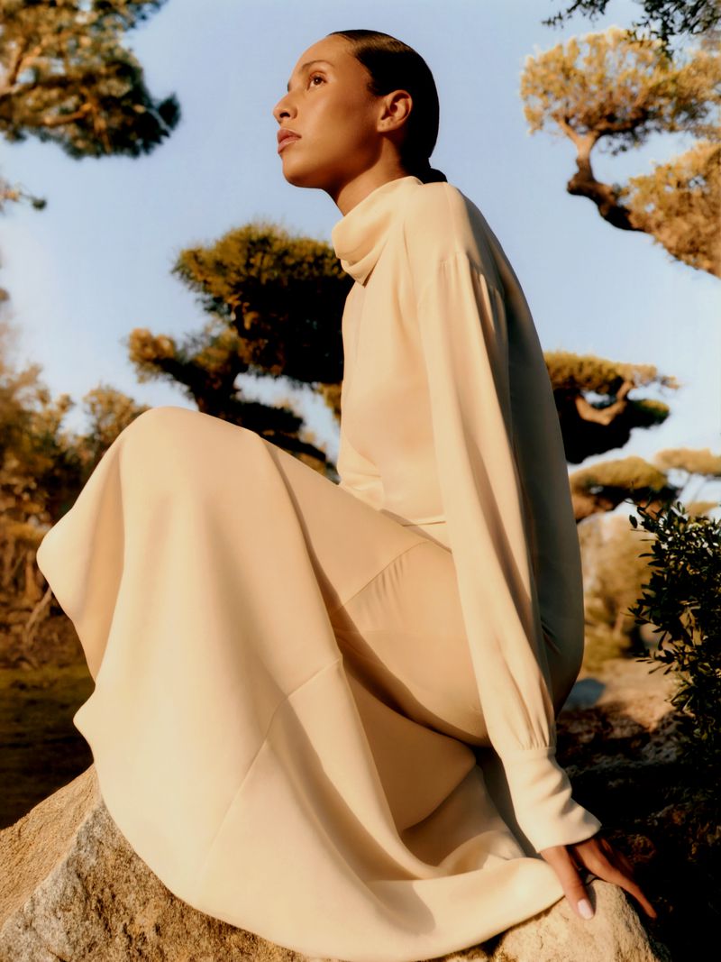 Adult female model wearing a beige dress sitting on a rock in an outdoor garden setting with ornamental trees and a bright sky.