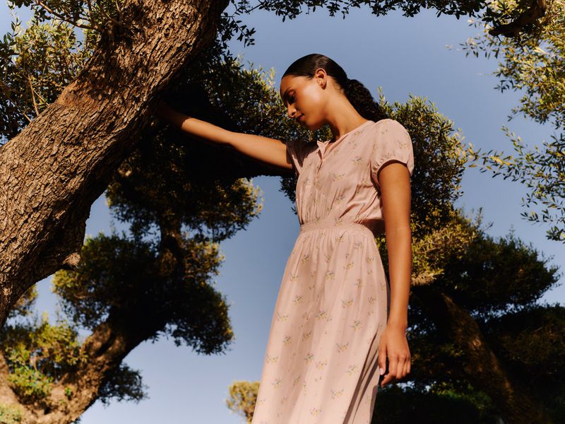 Woman in floral dress poses near tree against a blue sky. The scene captures natural light and a serene, outdoor fashion moment.