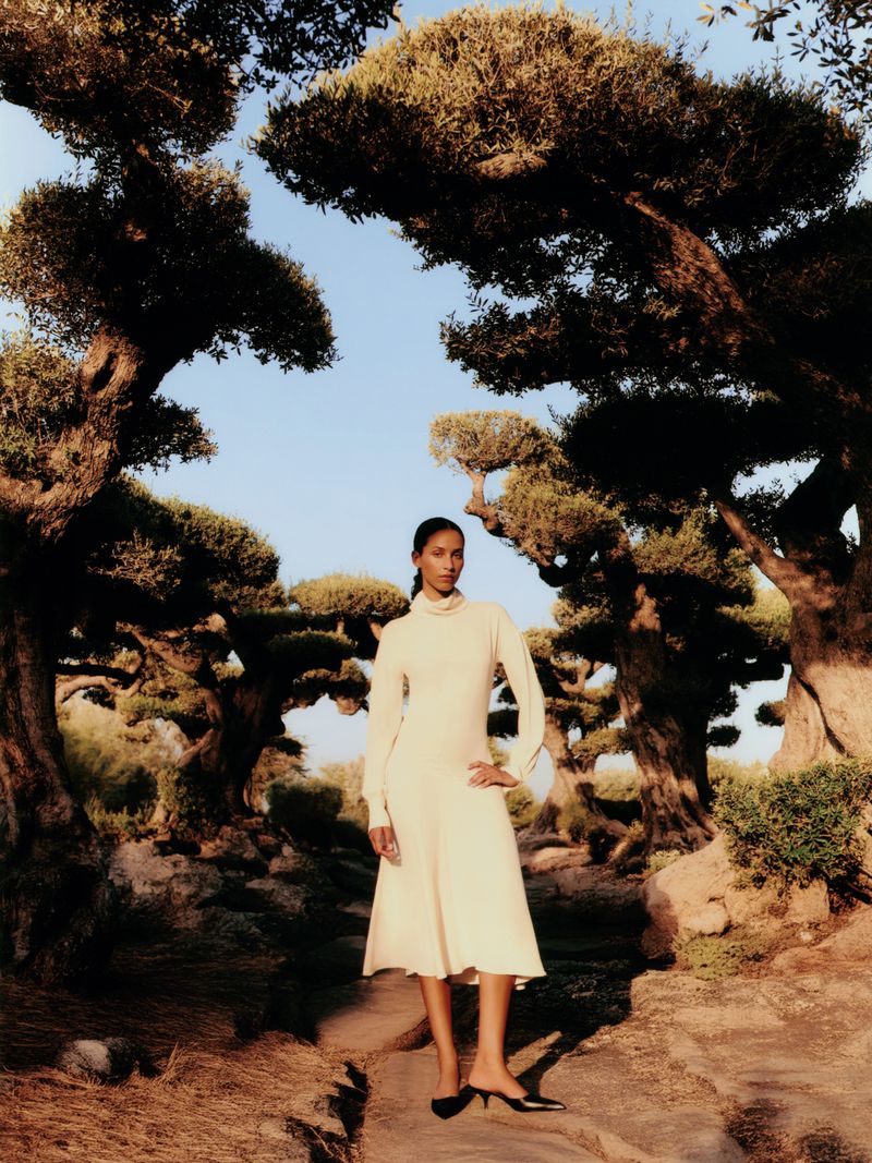 Woman poses in an off-white, long-sleeved dress and black shoes along a stone path surrounded by olive trees under a blue sky.
